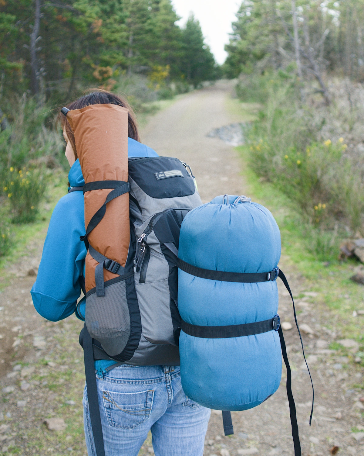 A woman wearing a backpack with a sleeping bag and sleeping pad strapped to it