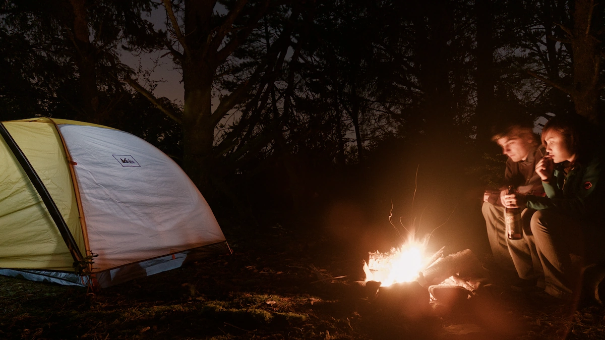 A man and women stare into a campfire with a lit up tent to their side