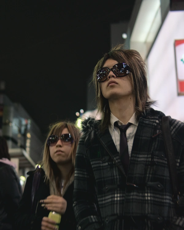 A Japanese couple wearing large sunglasses at night look at the city lights around them