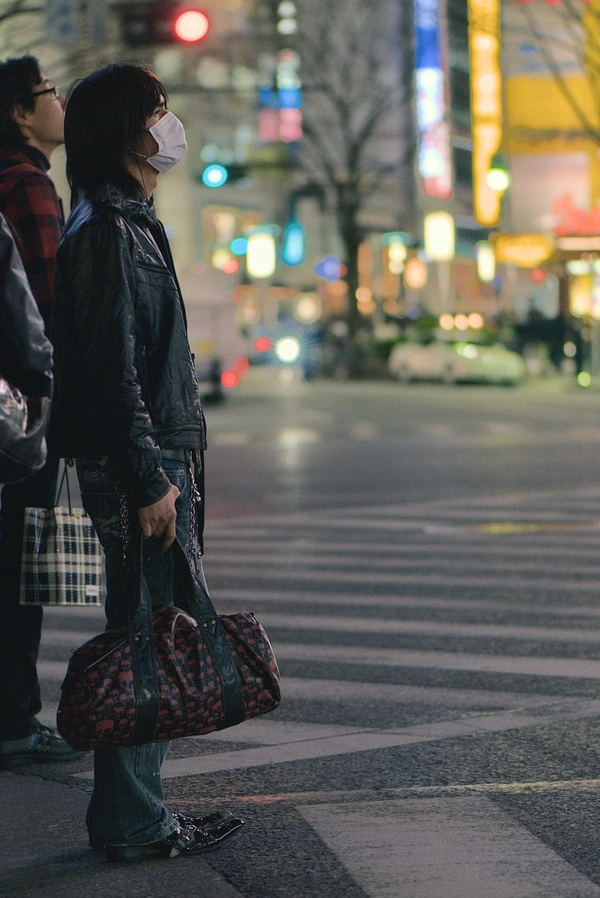 A man wearing a leather jacket and holding a leather bag waits on the curb to cross the street